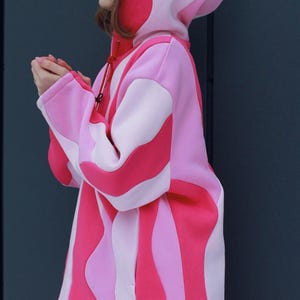 May include: A pink and red hooded sweatshirt with a unique wavy stripe design. The sweatshirt features a drawstring hood and long sleeves. The color palette includes shades of pink and red. The garment is made of a soft material.