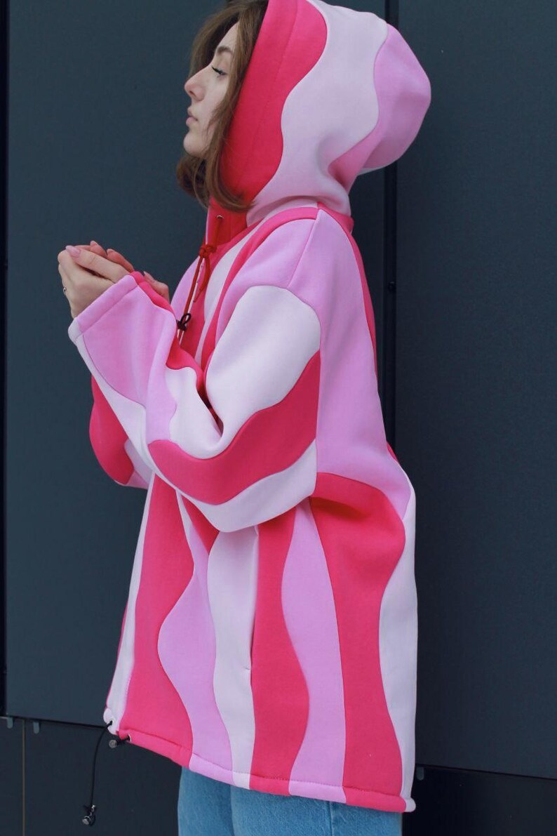 May include: A pink and red hooded sweatshirt with a unique wavy stripe design. The sweatshirt features a drawstring hood and long sleeves. The color palette includes shades of pink and red. The garment is made of a soft material.