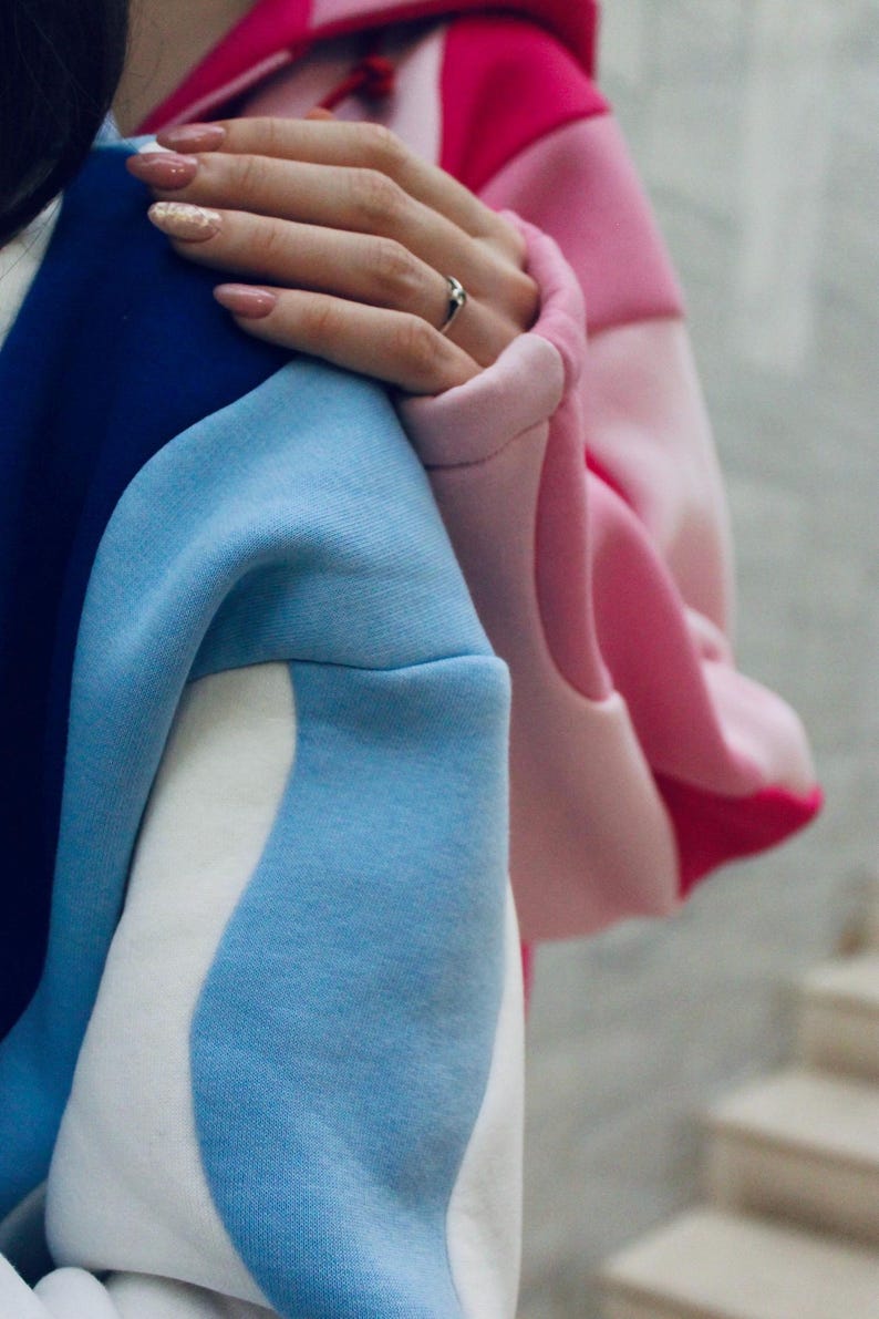 May include: A color-blocked hoodie in shades of blue, white, and pink. The design features a combination of solid and patterned sections. The sleeves are a light blue, and the hood is a gradient of pink hues. The fabric appears to be a soft, comfortable material.