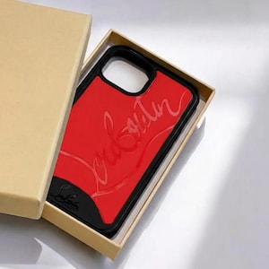 Könnte beinhalten: Eine rote Handyhülle mit dem Wort "Louboutin" in Schreibschrift auf der Oberfläche geprägt. Die Hülle hat einen schwarzen Rand und wird in einer beigefarbenen Box präsentiert. Die Handyhülle hat eine leuchtend rote Farbe.