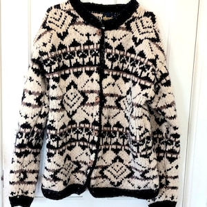 May include: A cream-colored, button-down cardigan sweater with a geometric pattern in black and brown. The sweater has a black collar, cuffs, and trim. The buttons are a light brown color. The sweater is hanging on a white hanger.