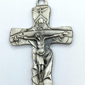 May include: A silver-colored crucifix pendant with a detailed depiction of Jesus Christ on the cross. The top of the cross features a dove and a depiction of God. The pendant has a loop for hanging.
