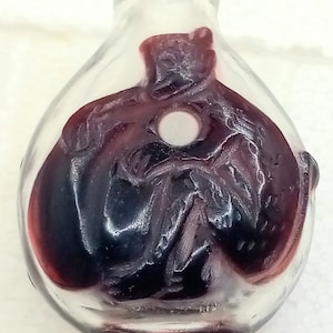May include: Clear glass bottle with a dark red, detailed interior design. The bottle has a rounded shape with a small opening at the top. The interior design appears to be a stylized figure or pattern. The bottle is likely decorative.