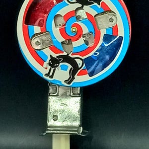May include: A vintage toy featuring a circular design with red, white, and blue concentric circles. Black cat illustrations are placed around the circle. The toy has a metal base and a cream-colored handle.