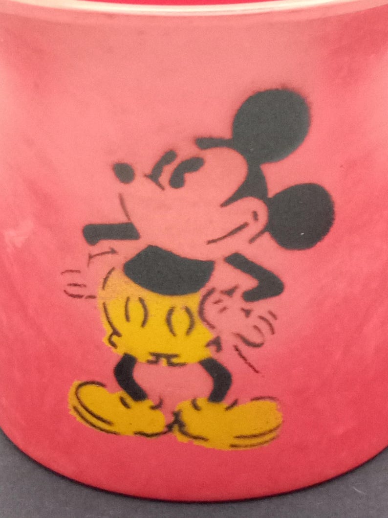 May include: Red ceramic mug with a black silhouette of Mickey Mouse. Mickey is wearing yellow shorts and black shoes.