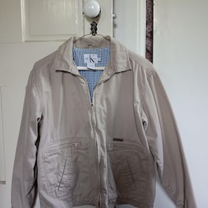 May include: A tan Calvin Klein jacket with a zip-up front, two front pockets, and a small logo on the chest. The jacket has a light blue and white checkered lining and is hanging on a white door.