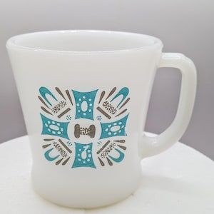 May include: White ceramic mug with a turquoise and brown geometric design. The mug has a curved handle and a flared shape. The design is centered on the front of the mug and features a symmetrical pattern.