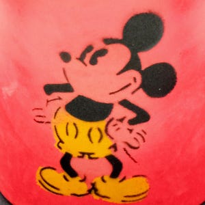 May include: A red ceramic mug with a black silhouette of Mickey Mouse. Mickey is wearing yellow shorts and is standing with his arms at his sides.
