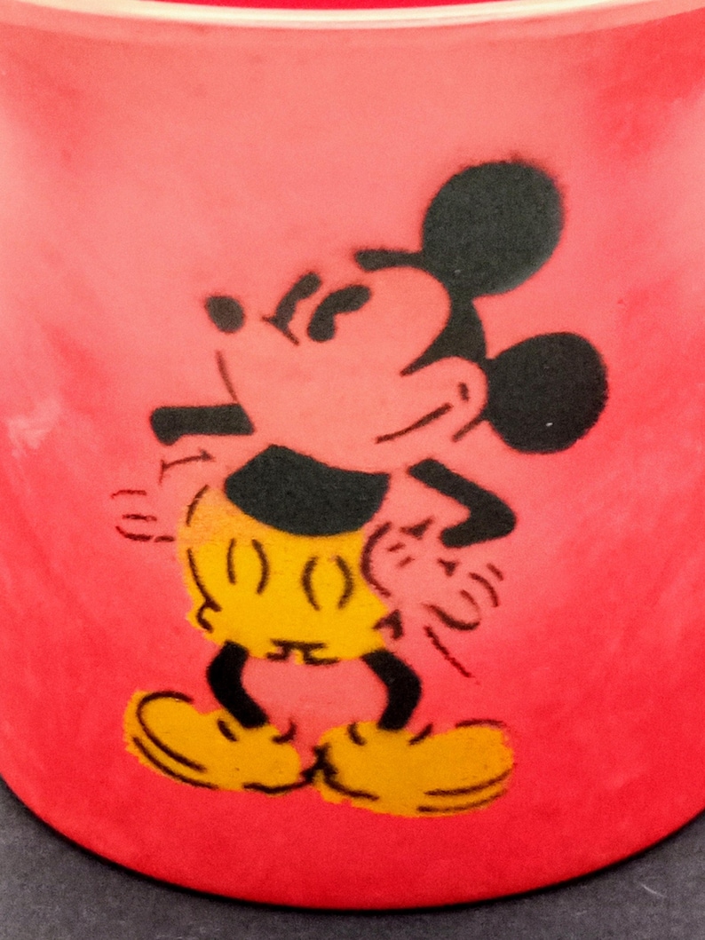 May include: A red ceramic mug with a black silhouette of Mickey Mouse. Mickey is wearing yellow shorts and is standing with his arms at his sides.
