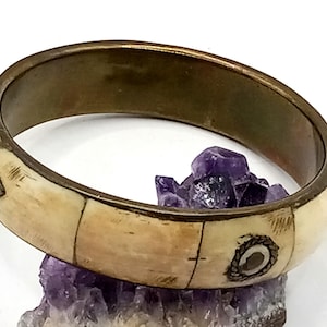 May include: Ivory-colored bangle bracelet with a brass-colored metal band. The bracelet features decorative metal accents and is segmented into rectangular sections. The bracelet is resting on a cluster of purple crystals.