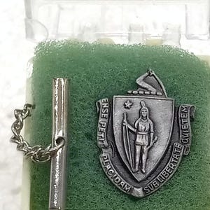May include: A silver-toned tie tack and emblem set. The emblem features a shield with a Native American figure and the words "ENSE PETIT PLACIDAM SUB LIBERTATE QUIETEM". The set is displayed in a clear plastic case with green foam.