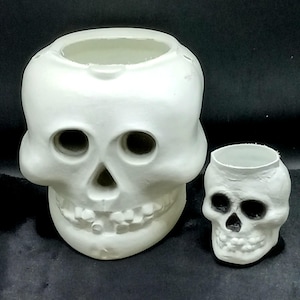 May include: A large white skull-shaped container with a hollow top and a smaller white skull-shaped container beside it. The smaller skull features dark, recessed eye sockets and a nasal cavity, while the larger skull has a smooth, matte finish.