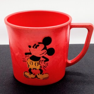 May include: Red plastic Mickey Mouse mug with a black and yellow image of Mickey Mouse on the front. The mug has a handle on the right side and the words "Frosty Ware" are printed on the handle.