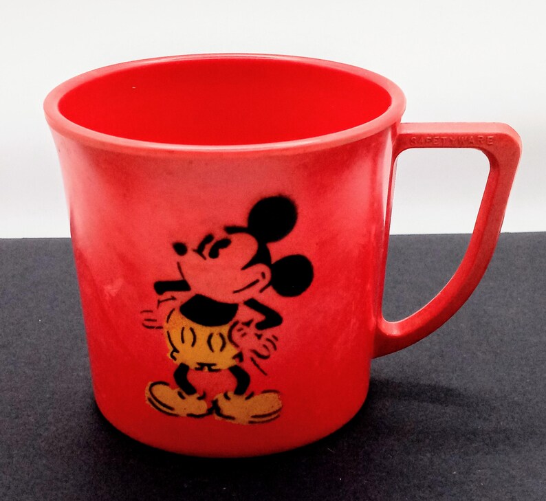 May include: Red plastic Mickey Mouse mug with a black and yellow image of Mickey Mouse on the front. The mug has a handle on the right side and the words "Frosty Ware" are printed on the handle.