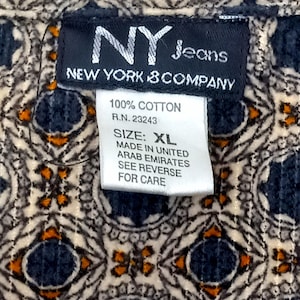 May include: Close-up of a patterned fabric with a label from NY Jeans, New York & Company. The label indicates 100% cotton, size XL, and made in the United Arab Emirates. The fabric has a repeating geometric design in navy, beige, and orange.