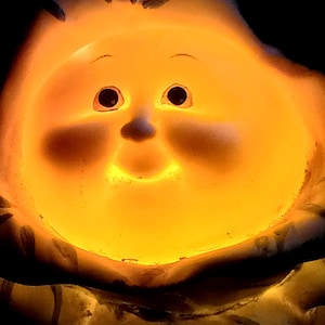 May include: A ceramic figurine of a round-faced character with a smooth surface, illuminated from within by a warm, glowing orange light. The face features dark, circular eyes, a small nose, and a subtle mouth, all highlighted by the internal glow.
