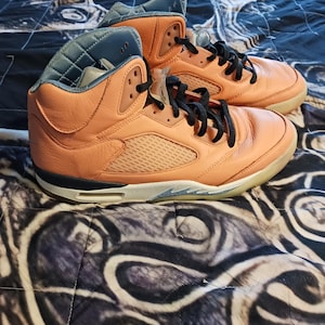 May include: A pair of orange and black high-top trainers. The trainers have a leather upper with black laces, a grey tongue, and a white and grey sole. The shoes are on a patterned bedspread.