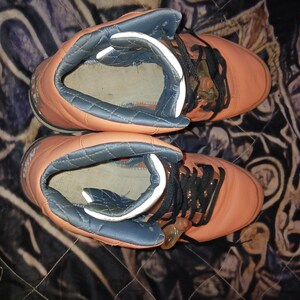 May include: Pair of orange high-top trainers with black laces. The trainers have a padded, blue-grey collar and a white interior. The shoes are set against a patterned background.