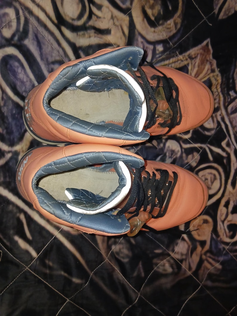 May include: Pair of orange high-top trainers with black laces. The trainers have a padded, blue-grey collar and a white interior. The shoes are set against a patterned background.