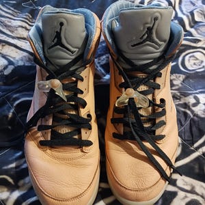 May include: Pair of peach-coloured high-top trainers with black laces and a grey tongue featuring a black Jumpman logo. The trainers have a light blue interior and clear lace locks. The shoes are displayed on a patterned surface.