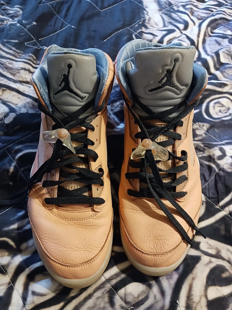 May include: Pair of peach-coloured high-top trainers with black laces and a grey tongue featuring a black Jumpman logo. The trainers have a light blue interior and clear lace locks. The shoes are displayed on a patterned surface.