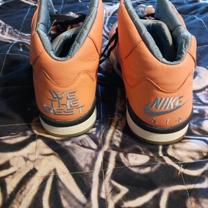 May include: Pair of orange and grey high-top trainers. The trainers have black laces and a white sole with a black stripe. The back of the left shoe says "WE THE BEST" and the right shoe says "NIKE AIR".