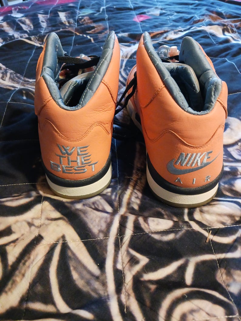 May include: Pair of orange and grey high-top trainers. The trainers have black laces and a white sole with a black stripe. The back of the left shoe says "WE THE BEST" and the right shoe says "NIKE AIR".