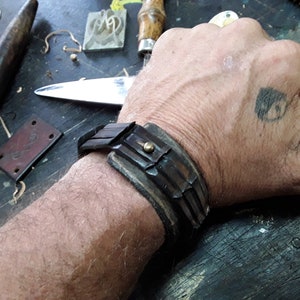 May include: A brown leather bracelet with a metal accent. The bracelet is worn on a person's wrist.