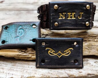 May include: Three leather labels with different designs. The top label is black with gold lettering that reads "NHJ" and has gold rivets. The middle label is blue with a gold design. The bottom label is black with a gold design.