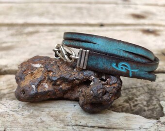 May include: A turquoise leather bracelet with a silver clasp and a turquoise design. The bracelet is wrapped around a rock.
