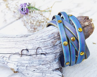 May include: A stack of blue leather bracelets with gold-colored studs. The bracelets are wrapped around a piece of driftwood. The background includes white and purple flowers, creating a natural, bohemian aesthetic.
