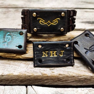 May include: Four leather patches with different designs. One patch is black with gold lettering that says "NHJ". Another patch is black with a gold design. The third patch is brown with a turquoise design. The fourth patch is brown with a gray design.