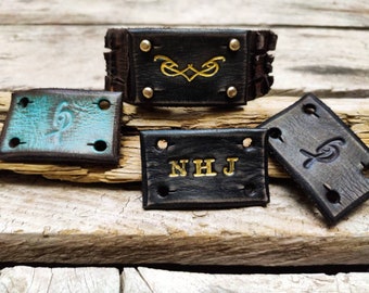 May include: Four leather patches with different designs. One patch is black with gold lettering that says "NHJ". Another patch is black with a gold design. The third patch is brown with a turquoise design. The fourth patch is brown with a gray design.
