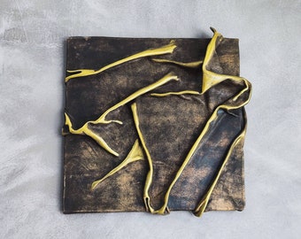 May include: A square canvas with a textured, dark brown background. The canvas is decorated with gold paint in a flowing, abstract design.