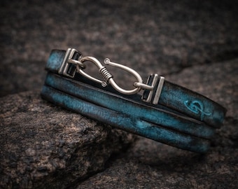 May include: A handcrafted leather bracelet with a distressed turquoise finish. The bracelet features a silver-toned hook clasp and is composed of three leather bands. The bracelet is resting on a dark, textured surface.
