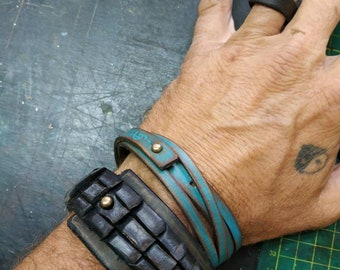 Op de afbeelding: Twee leren armbanden om de pols van een persoon. De bovenste armband is een dunne, turquoise leren band met een gouden accent. De onderste armband is een dikke, zwarte leren armband met een textuur en een gouden accent.
