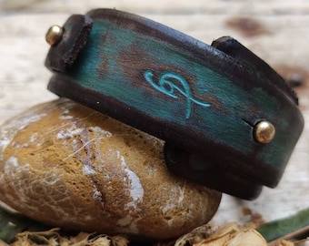 May include: A brown leather bracelet with a turquoise blue design and gold accents. The bracelet is on a rock.