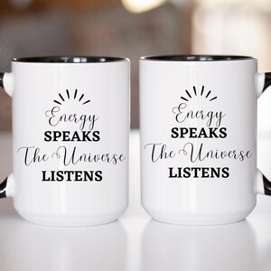 May include: Two white ceramic mugs with black interiors and handles. Each mug features the text "Energy Speaks, The Universe Listens" in black script and a sunburst design above the text. The mugs are on a white surface.