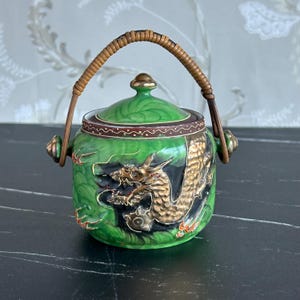 May include: A green ceramic container with a lid and a woven handle. The container features a gold dragon design and red accents. The lid is green with a gold finial. The handle is made of woven brown material.