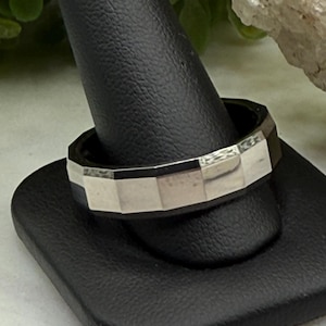 May include: A silver-colored ring with a geometric, faceted design. The ring is displayed on a black ring holder. The ring's surface is divided into rectangular segments, creating a modern and angular aesthetic. The ring is likely made of metal.
