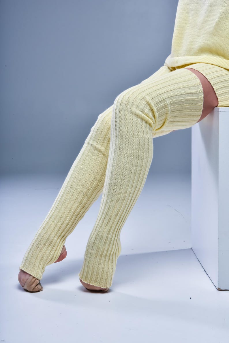 May include: Pale yellow ribbed knit leg warmers. The leg warmers are long, extending from the foot to the upper thigh. The ribbed texture is visible. The person is wearing a matching pale yellow sweater.