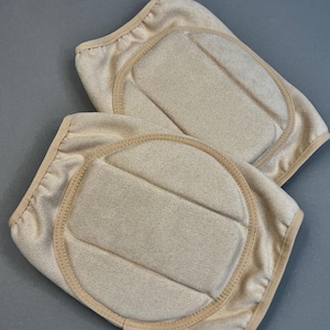 Può includere: Due ginocchiere beige con un design imbottito. I cuscinetti hanno una forma arrotondata con un centro imbottito rettangolare e sono bordati con un bordo beige più chiaro. Lo sfondo è grigio.