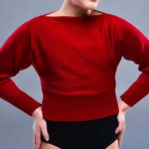 May include: A red sweater with a wide neckline and long sleeves. The sweater has a ribbed hem and cuffs. The model is wearing black bottoms. The sweater is a solid color and appears to be made of a soft knit material.
