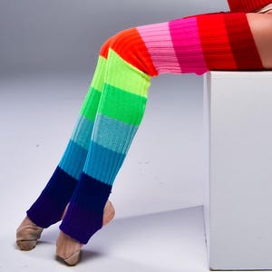 May include: Rainbow striped leg warmers with a purple ombre at the bottom. The leg warmers are worn on a person's legs.