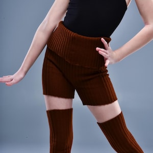 May include: A brown ribbed knit shorts set with matching leg warmers. The shorts have a wide, folded waistband. The outfit is worn with a black vest top. The ribbed knit fabric has a vertical pattern.