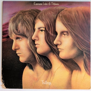 May include: Album cover art featuring three side profiles of individuals with long hair, set against a purple and brown background. The text "Emerson Lake & Palmer" is at the top, and "Trilogy" is at the bottom.