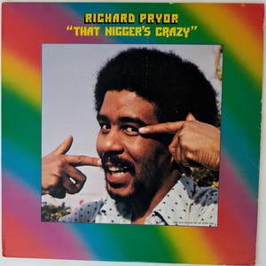 May include: Album cover for Richard Pryor's comedy album "That Nigger's Crazy." The cover features a photo of Pryor pointing at his eye and mouth, with a rainbow-colored border. The album title is in yellow text.