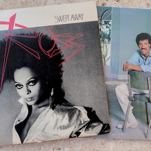 May include: Two vinyl record album covers. The left cover features Diana Ross with the text "DIANA ROSS" and "SWEPT AWAY" in red and black. The right cover shows a man in a light blue shirt and white pants sitting on a metal chair.