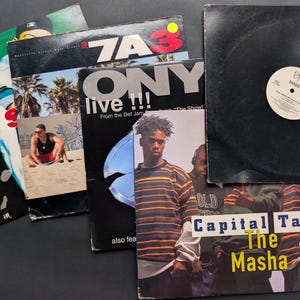 May include: A collection of vinyl record album covers, featuring various artists and album titles. The covers display diverse graphics, including images of people, text, and abstract designs. The records are stacked on a dark surface.