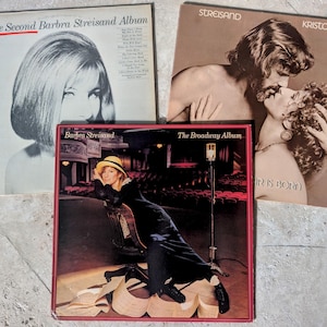 May include: Three vinyl record albums featuring Barbra Streisand. One cover shows a black and white portrait titled The Second Barbra Streisand Album. Another is the A Star Is Born soundtrack. The third is The Broadway Album with a color photo.